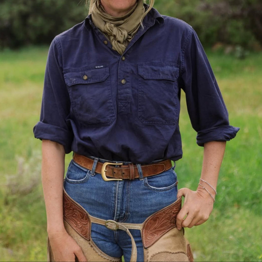 Person wearing a blue shirt, cowboy hat, and jeans standing in a grassy field with trees in the background.